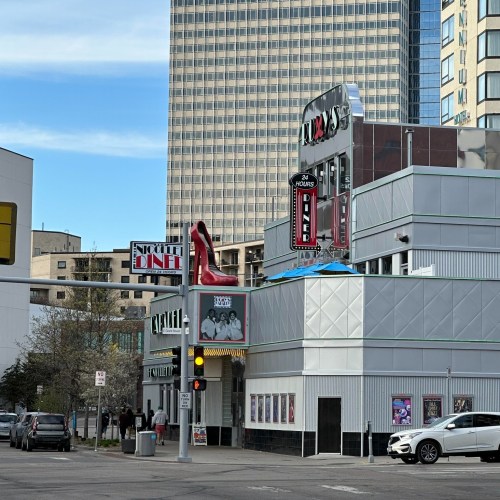 A diner with a large red high heel sign, surrounded by modern buildings and parked cars on a city street.