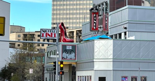 A large red high heel shoe atop a diner sign, with a 24-hour sign visible nearby. Urban setting with buildings in the background.