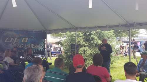A speaker presents under a tent at a UFO and paranormal event, with an audience seated in front.