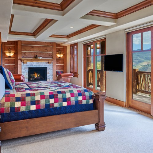 Cozy bedroom with a quilted bed, fireplace, and large windows showcasing a scenic view.