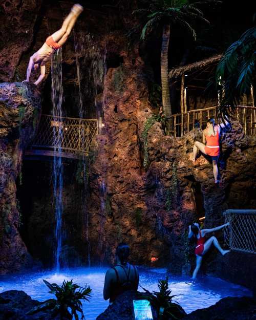 A person dives into a pool from a rocky ledge while others climb and explore the tropical-themed environment.