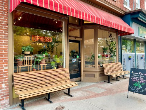 A cozy café with a red awning, open sign, and benches outside, surrounded by plants and a chalkboard menu.