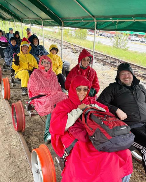 A group of people in colorful rain gear sitting on a train-like cart under a green canopy, smiling and enjoying the ride.