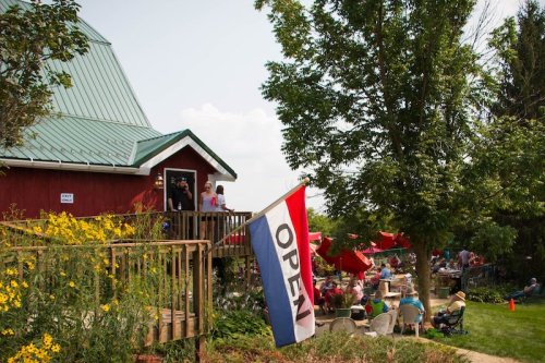 A red barn with a green roof, featuring an "Open" flag, surrounded by people enjoying the outdoor seating area.