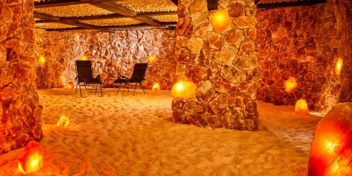A tranquil salt room with stone walls, salt lamps, and two lounge chairs on a sandy floor, creating a calming atmosphere.