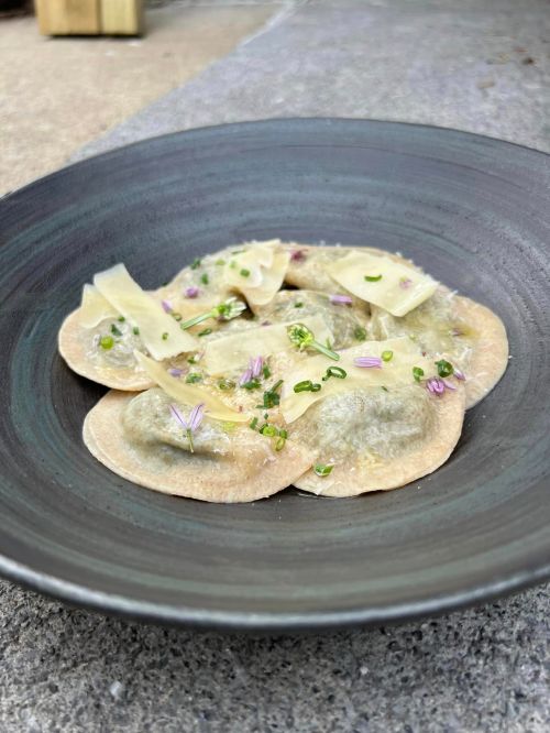A plate of ravioli topped with herbs and cheese, served in a dark bowl on a textured surface.