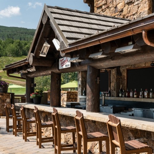 A rustic outdoor bar with wooden stools, stone walls, and a scenic view of greenery and mountains in the background.