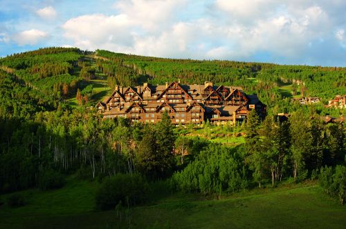 A large, rustic lodge nestled among lush green hills and trees under a partly cloudy sky.