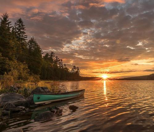 A serene lake at sunset, with a canoe on the shore and vibrant clouds reflecting on the water.