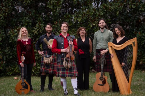 A group of six musicians in traditional attire, holding various instruments, standing outdoors in front of greenery.