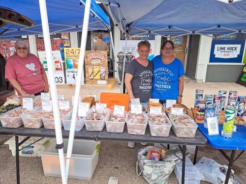 A market stall with three women smiling behind tables filled with packaged food items and drinks under a blue canopy.