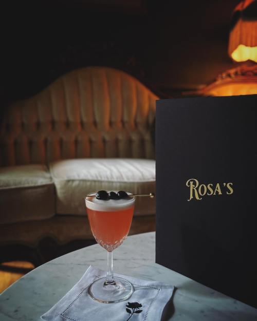 A cocktail with a frothy top and blackberries sits on a marble table next to a menu, with a cozy sofa in the background.