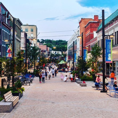 A lively street scene with people walking, shops lining the sides, and colorful banners under a blue sky.