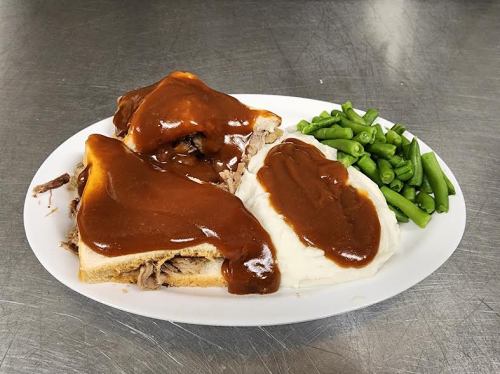 A plate with two sandwiches topped with brown gravy, mashed potatoes, and a side of green beans.