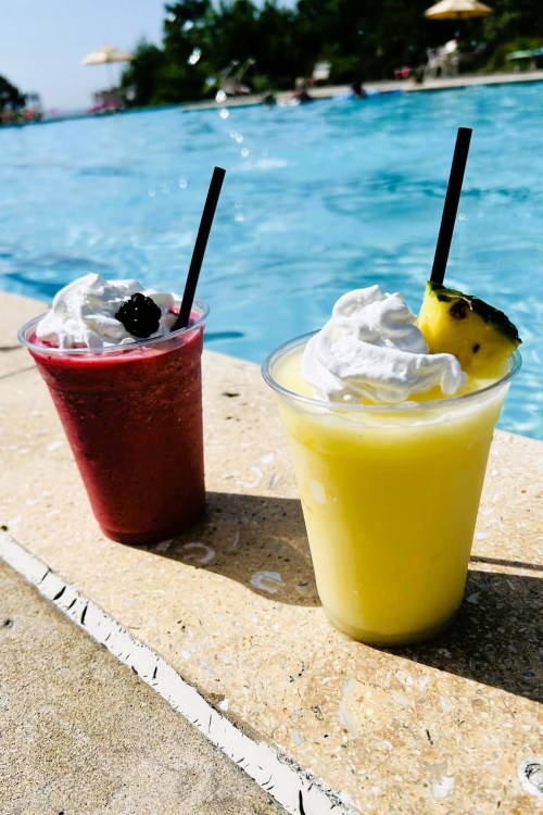 Two colorful drinks with whipped cream and fruit garnishes sit by a sparkling blue pool.