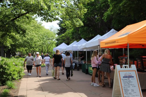 A bustling outdoor market with people walking among white and orange tents, surrounded by lush greenery.