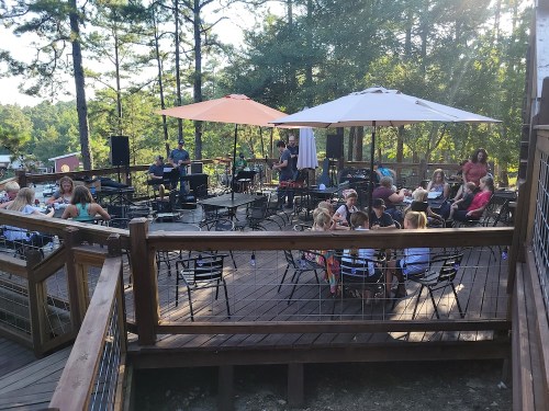 A lively outdoor gathering on a wooden deck with people sitting at tables under umbrellas, enjoying music and conversation.