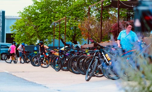 A row of parked bicycles along a pathway, with people walking and greenery in the background.