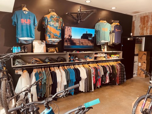 A bike shop interior featuring clothing displays, helmets, and a TV screen on the wall. Bikes are visible in the foreground.