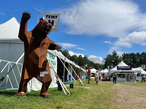 A large wooden bear sculpture stands near festival tents under a blue sky with scattered clouds.