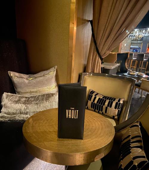A stylish lounge setting featuring a menu titled "The Vault" on a round table, with plush seating and elegant decor.