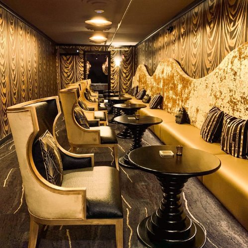 A stylish lounge with plush seating, gold accents, and elegant decor, featuring dark walls and intimate lighting.