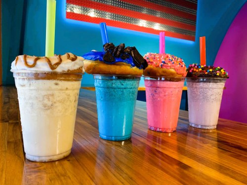 Four colorful milkshakes topped with donuts and sprinkles, served in clear cups on a wooden table.