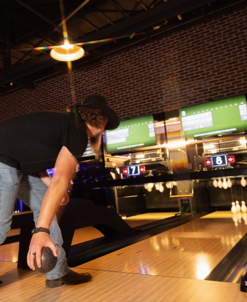 A person in a black shirt and hat prepares to bowl, with illuminated scoreboards in the background.