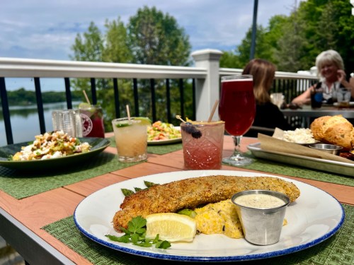 A plate of fried fish with a side of corn, drinks, and salads on a table overlooking a scenic lake.