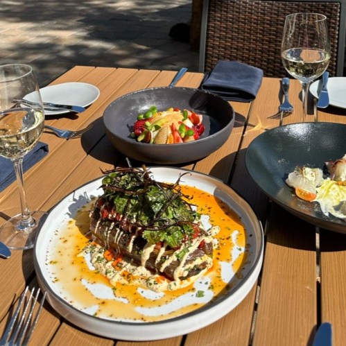A beautifully plated meal featuring layered meat, sauce, and greens, with a side of fruit salad and glasses of white wine.