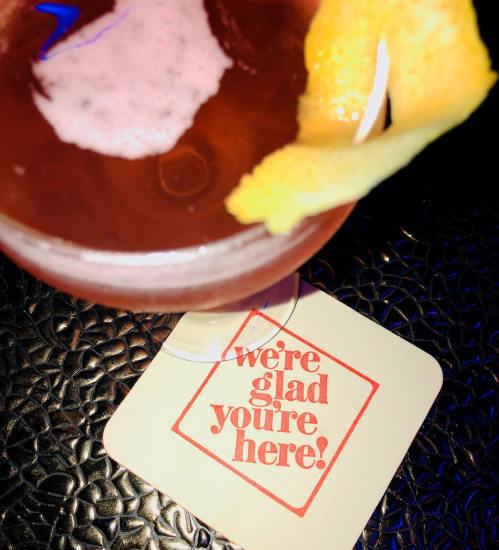 A cocktail with a lemon twist beside a coaster that says, "we're glad you're here!" on a textured surface.