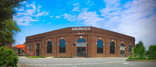 A brick building with large windows, labeled "Georgia Beer Co," set against a blue sky with scattered clouds.