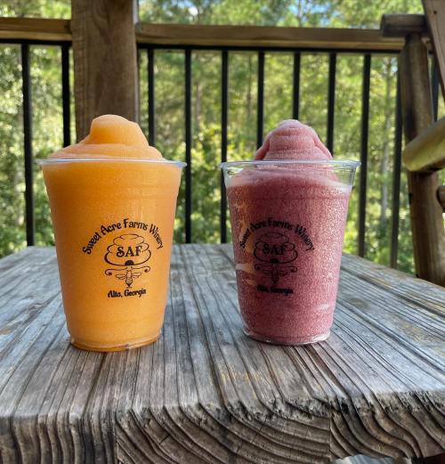Two colorful frozen drinks in clear cups, one orange and one purple, sitting on a wooden table outdoors.