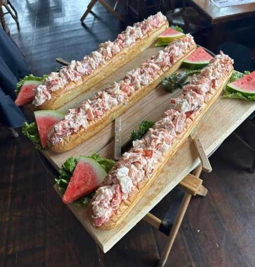 Three large sandwiches filled with seafood salad, garnished with lettuce and slices of watermelon on a wooden board.