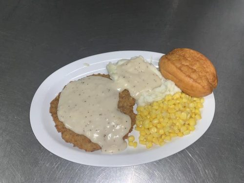 A plate with fried chicken topped with gravy, corn, mashed potatoes, and a biscuit.