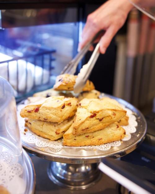 A hand using tongs to pick up scones from a silver platter in a bakery display.