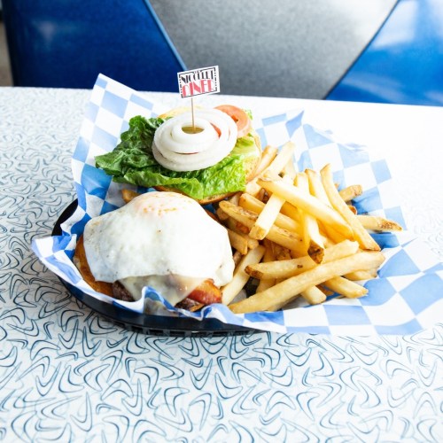 A plate of a burger topped with lettuce, tomato, onion, and a fried egg, served with a side of golden fries.