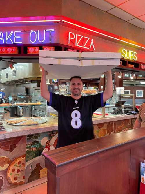 A man in a sports jersey holds a large pizza box in a colorful restaurant with neon signs.