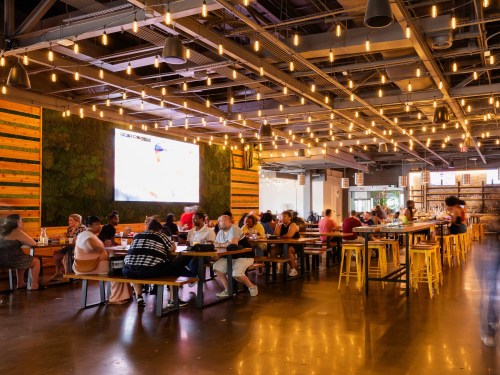 A lively restaurant interior with string lights, featuring diners at long tables and a large screen on the wall.