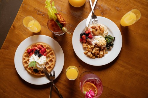 A wooden table with two plates of brunch: waffles topped with berries and cream, and a savory dish with shrimp and potatoes, plus drinks.