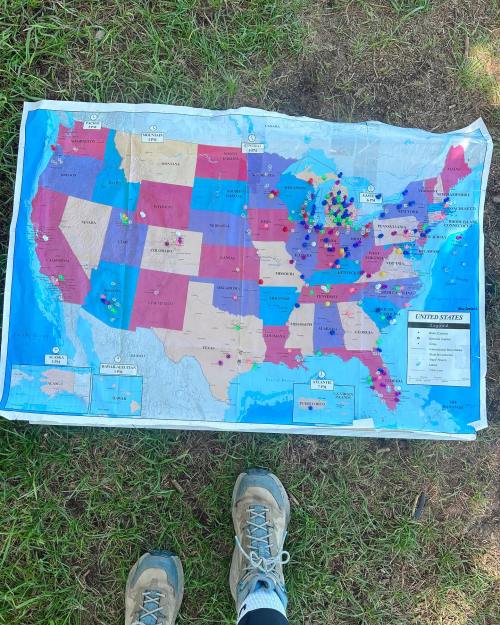 A map of the United States laid on grass, marked with colorful pins, with a pair of shoes visible at the bottom.
