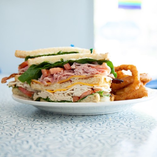 A tall sandwich stacked with turkey, ham, lettuce, and tomato, served with onion rings on the side.
