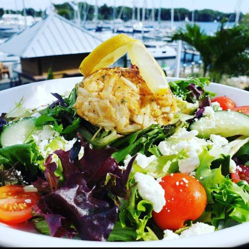 A vibrant salad topped with crab meat, lemon, and feta cheese, with a marina view in the background.