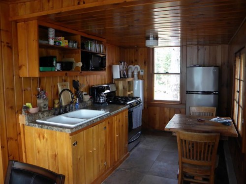 Cozy wooden kitchen with a sink, stove, fridge, and a dining table by a window, featuring rustic decor.