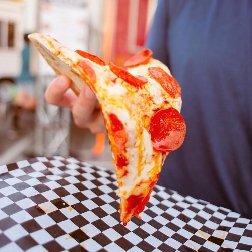 A hand holding a slice of pepperoni pizza over a checkered paper tray.