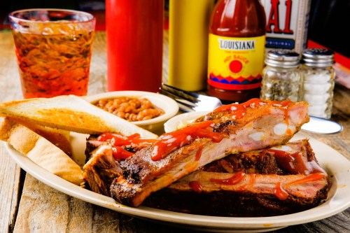 A plate of BBQ ribs with ketchup, toast, baked beans, and a glass of iced tea on a wooden table.