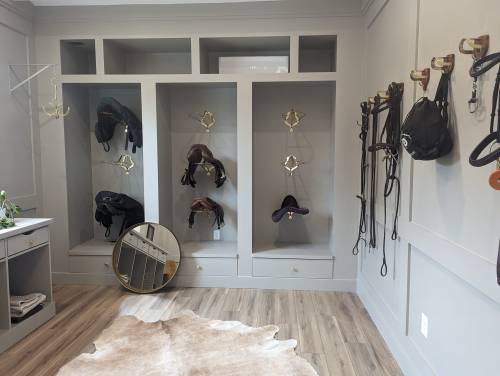 A stylish equestrian room featuring saddles, bridles, and a mirror against a gray wall with wooden flooring.