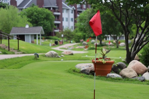 A red flag marks a golf hole on a lush green course, with trees and rocks in the background.