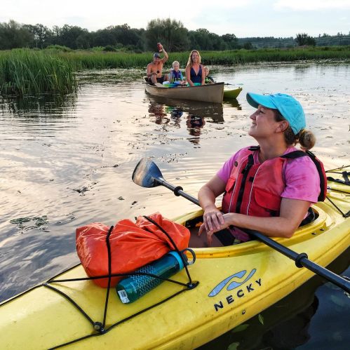 A woman in a kayak smiles while paddling, with a family in a canoe in the background on a calm waterway.