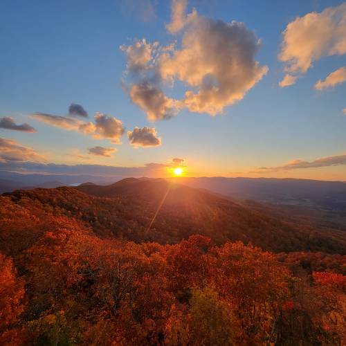 A vibrant sunset over rolling hills, surrounded by autumn foliage and a colorful sky with scattered clouds.